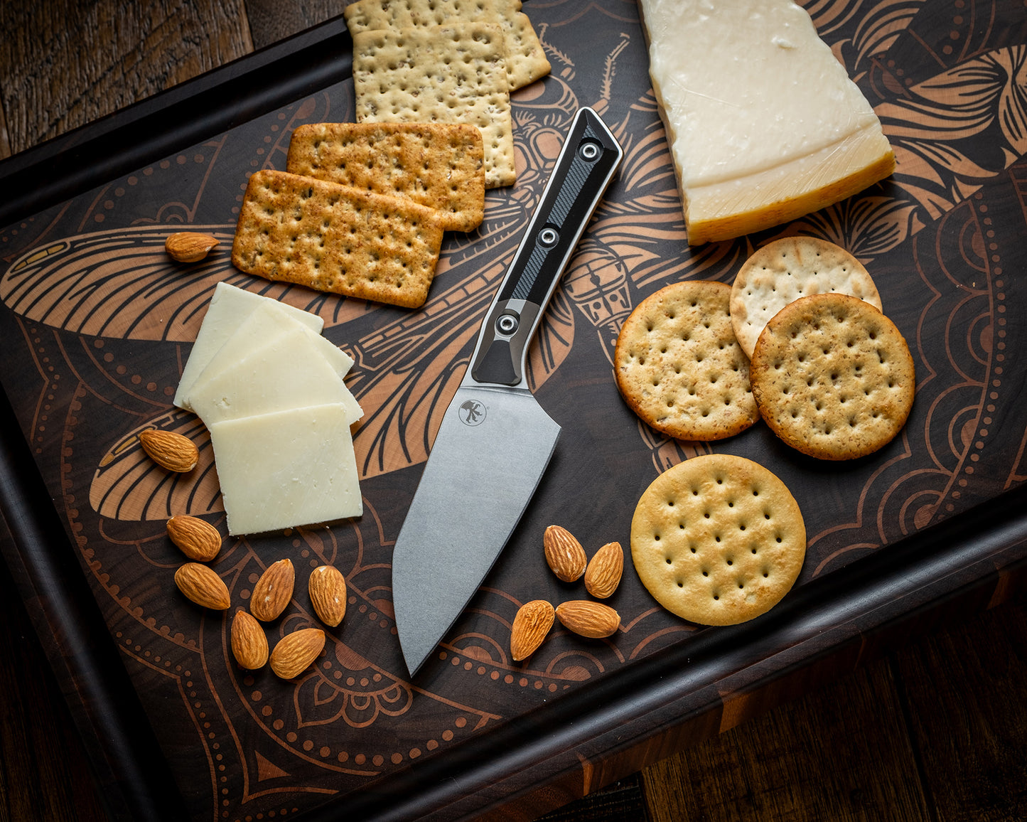 Microtech Culinary Santoku 4.2" Knife with black G10 handle, titanium bolsters and stonewashed stainless steel blade on a dragon fly cutting board surrounded by cheeses, crackers and nuts. Premium professional kitchen knife for precision chopping and modern cooking.