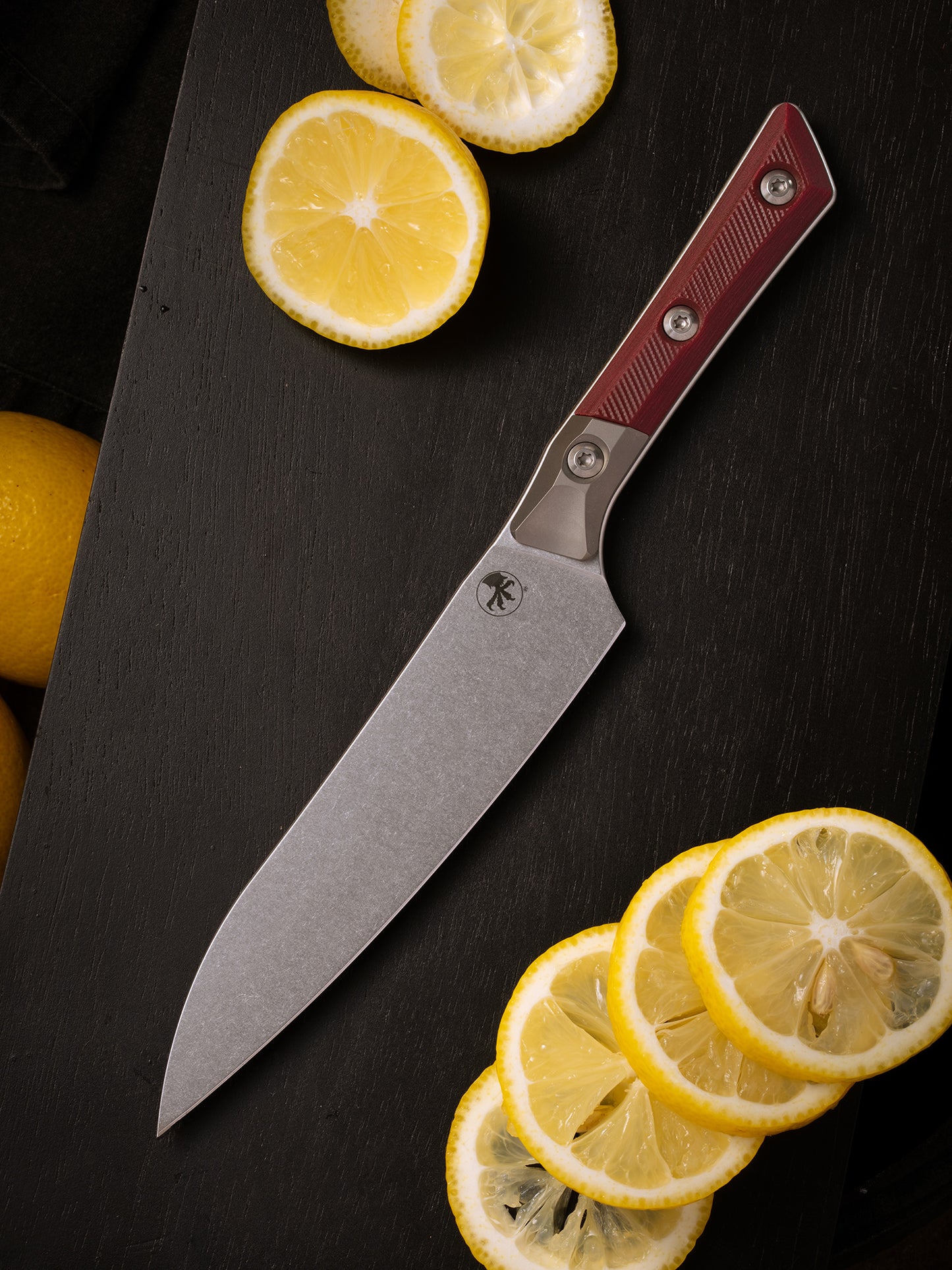 Microtech Culinary Utility Knife with merlot G10 handle, titanium bolsters and stonewashed stainless steel blade on a wooden cutting board surrounded by lemons. Premium professional kitchen knife for precision chopping and modern cooking.