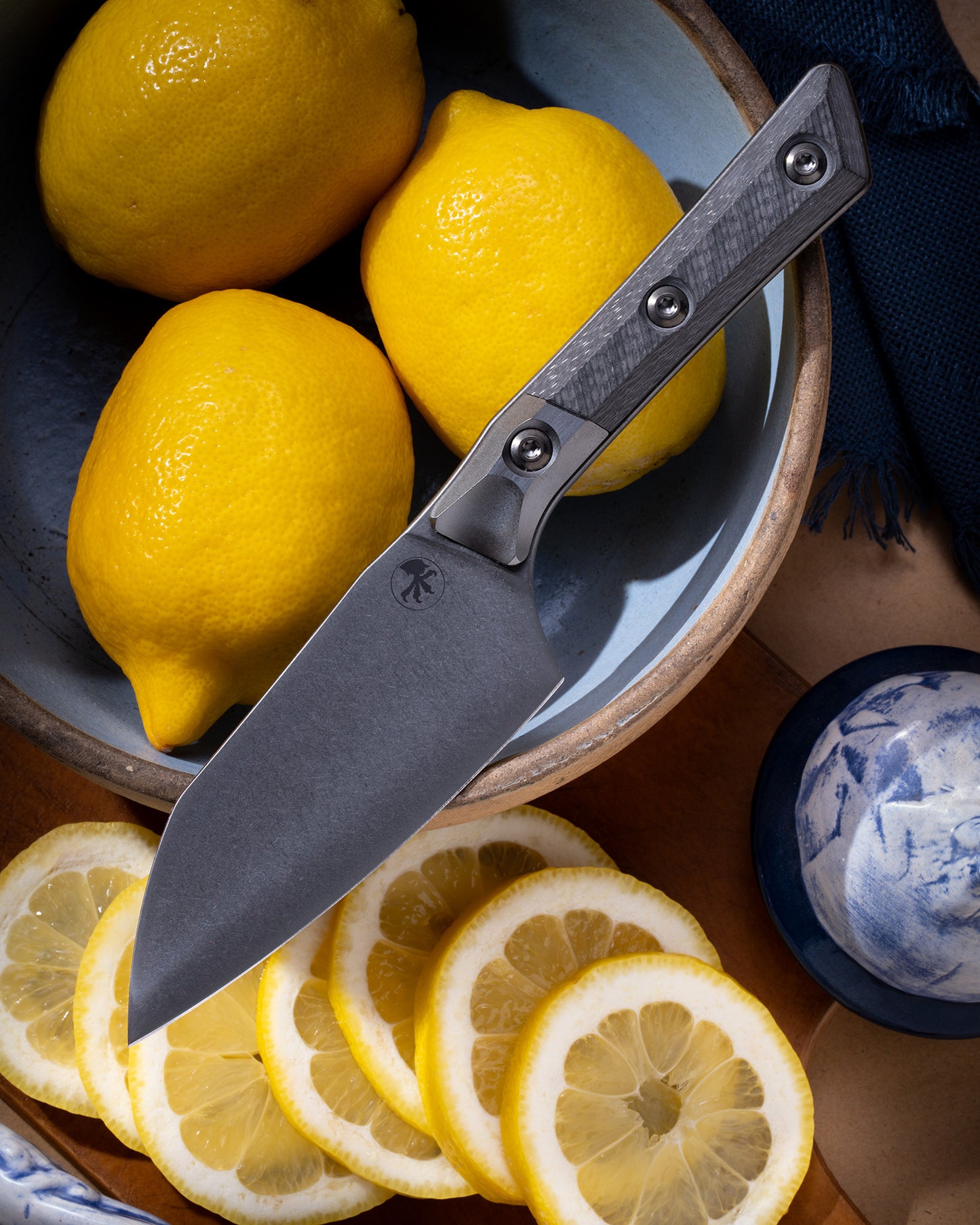 Microtech Culinary Santoku 4.2" Knife with carbon fiber handle, titanium DLC bolsters and DLC stainless steel blade on a bowl of lemons surrounded by cut lemons. Premium professional kitchen knife for precision chopping and modern cooking.