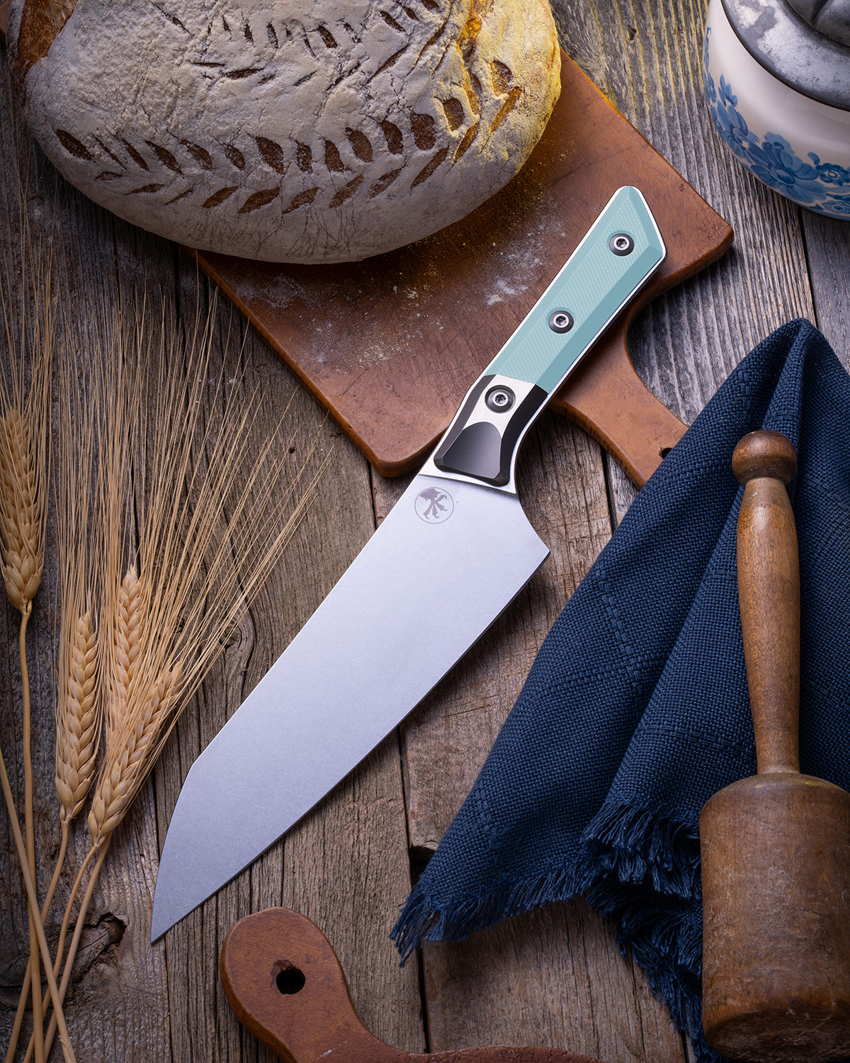 Microtech Culinary Chef Knife featuring a Caribbean Blue G10 handle and titanium bolster, displayed on a rustic wooden surface beside artisan bread, wheat stalks, and kitchen tools. Premium handcrafted chef knife for professional and home chefs.