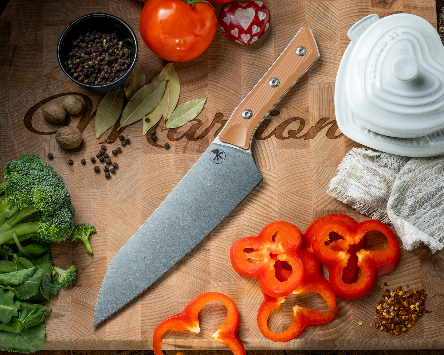 Microtech Culinary Chef Knife with ember G10 handle and stonewashed stainless steel blade on a wooden cutting board surrounded by fresh vegetables and spices. Premium professional kitchen knife for precision chopping and modern cooking.