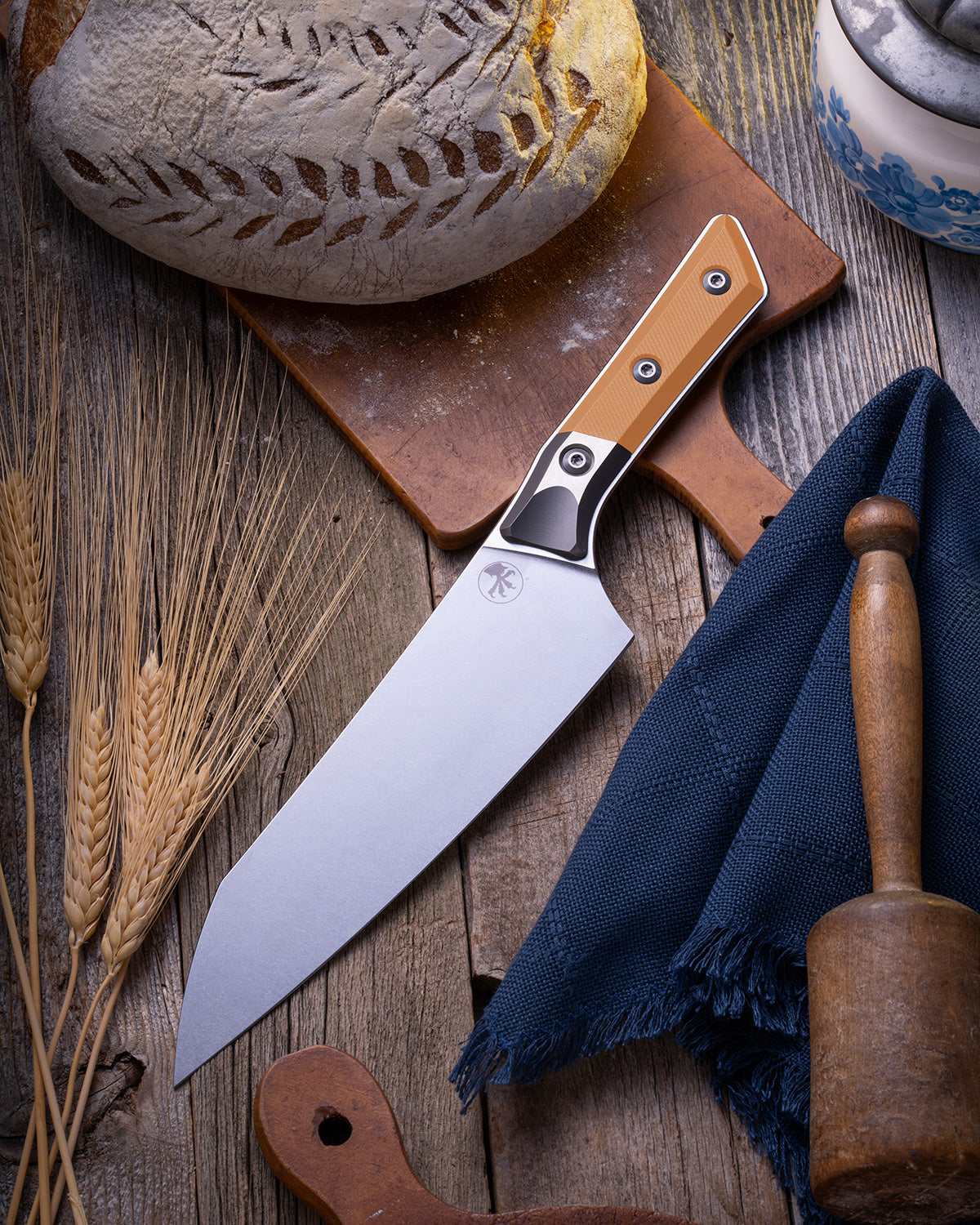 Microtech Culinary Chef Knife featuring an ember G10 handle and titanium bolster, displayed on a rustic wooden surface beside artisan bread, wheat stalks, and kitchen tools. Premium handcrafted chef knife for professional and home chefs.