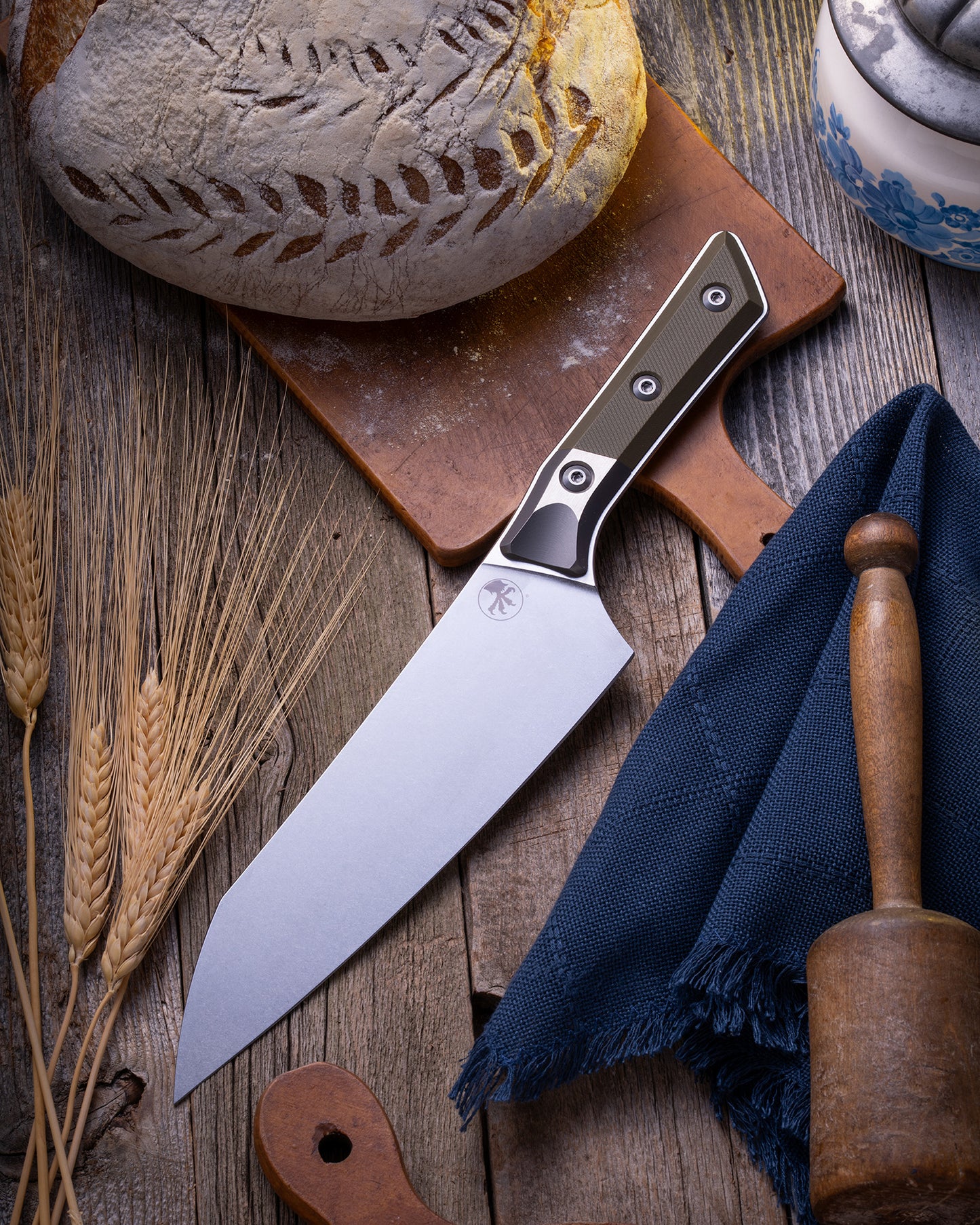 Microtech Culinary Chef Knife featuring an OD Green G10 handle and titanium bolster, displayed on a rustic wooden surface beside artisan bread, wheat stalks, and kitchen tools. Premium handcrafted chef knife for professional and home chefs.