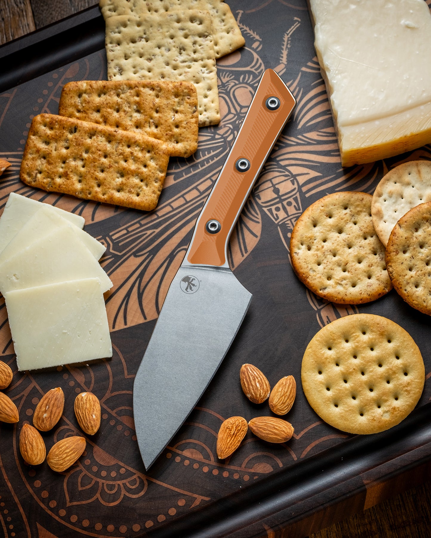 Microtech Culinary Santoku 4.2" Knife with ember G10 handle and stonewashed stainless steel blade on a dragon fly cutting board surrounded by cheeses, crackers and nuts. Premium professional kitchen knife for precision chopping and modern cooking.