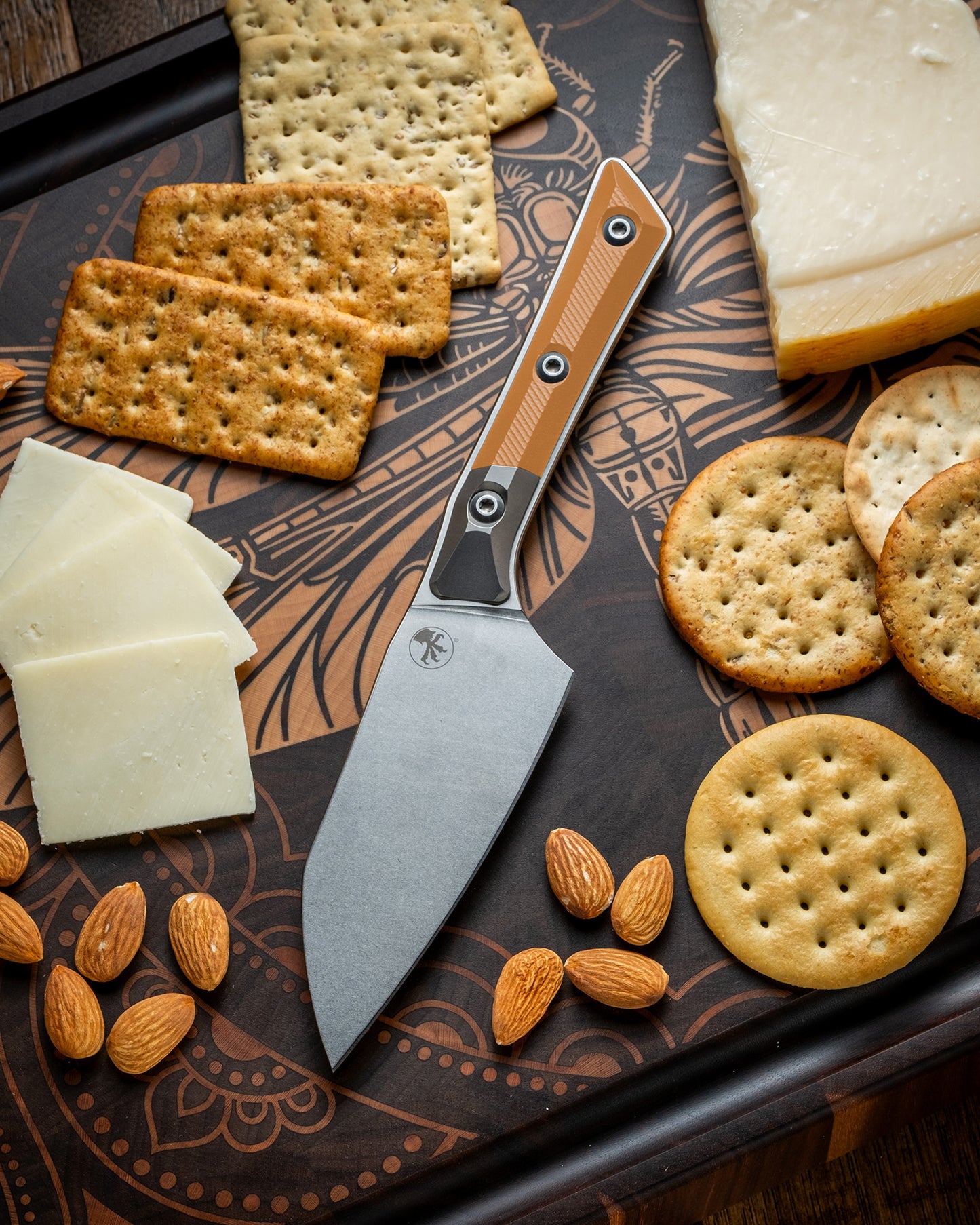 Microtech Culinary Santoku 4.2" Knife with ember G10 handle, titanium bolsters and stonewashed stainless steel blade on a dragon fly cutting board surrounded by cheeses, crackers and nuts. Premium professional kitchen knife for precision chopping and modern cooking.