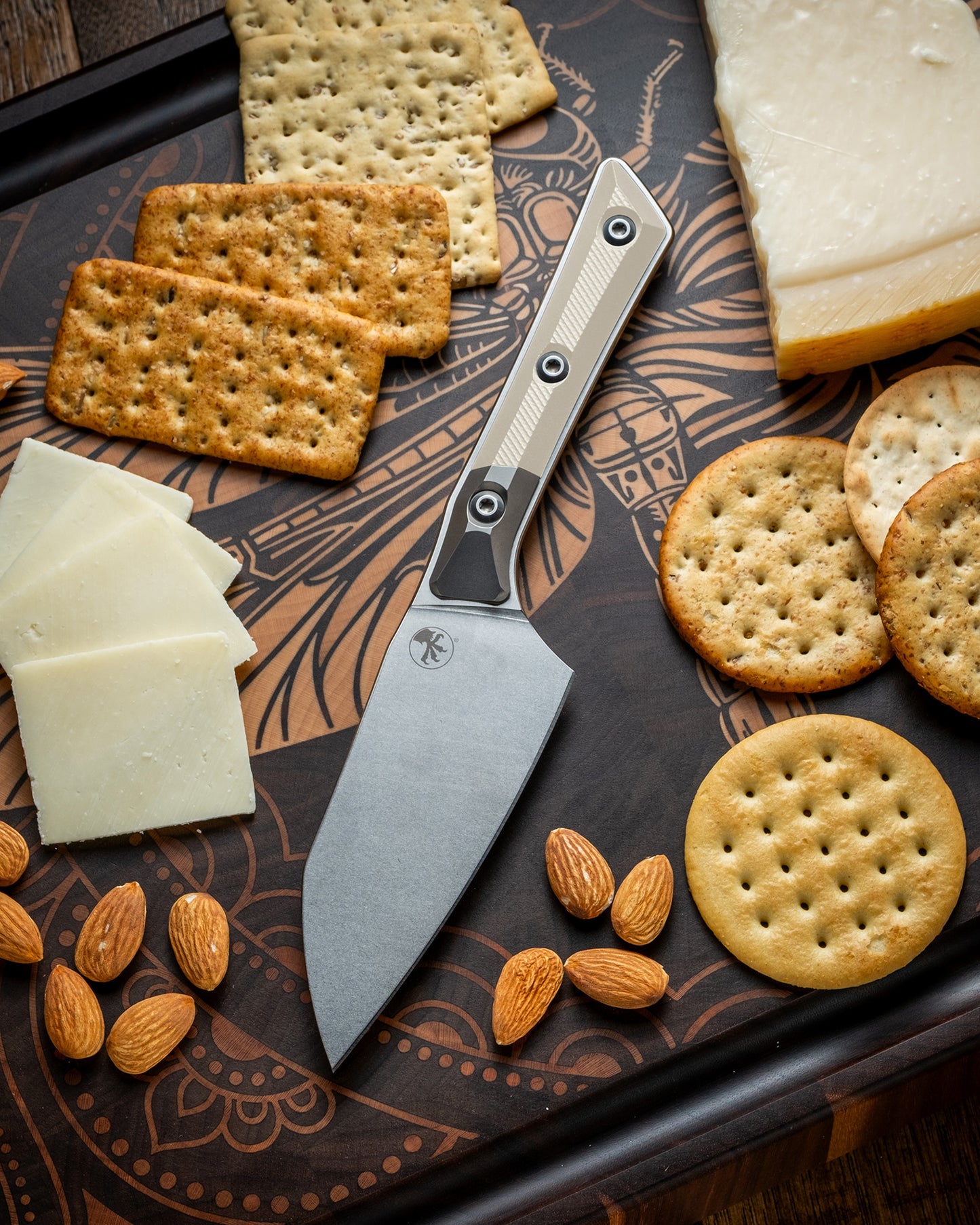Microtech Culinary Santoku 4.2" Knife with ivory G10 handle, titanium bolsters and stonewashed stainless steel blade on a dragon fly cutting board surrounded by cheeses, crackers and nuts. Premium professional kitchen knife for precision chopping and modern cooking.