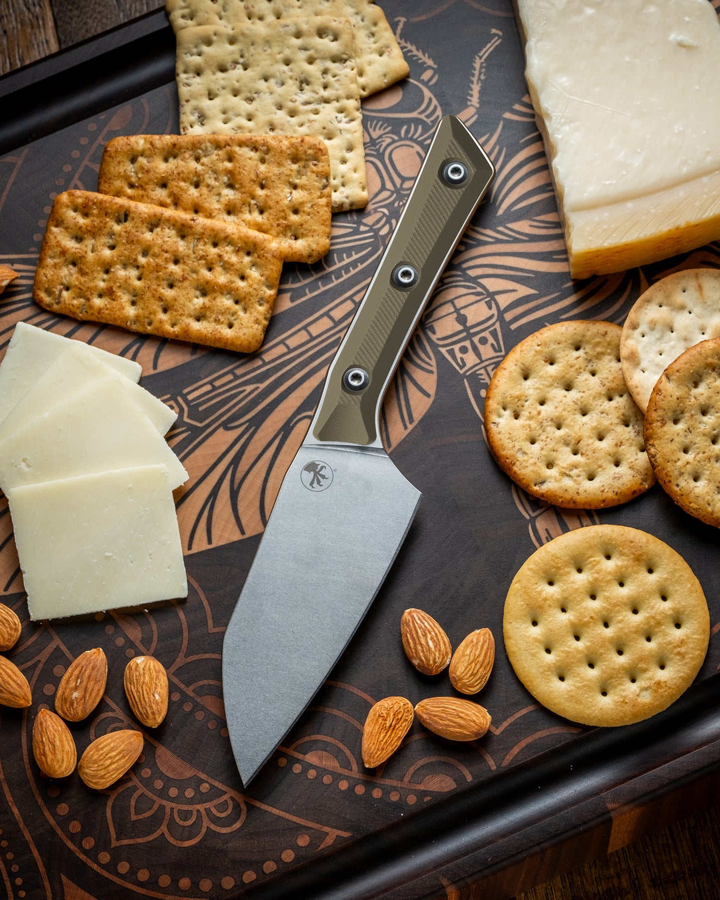 Microtech Culinary Santoku 4.2" Knife with OD green G10 handle and stonewashed stainless steel blade on a dragon fly cutting board surrounded by cheeses, crackers and nuts. Premium professional kitchen knife for precision chopping and modern cooking.