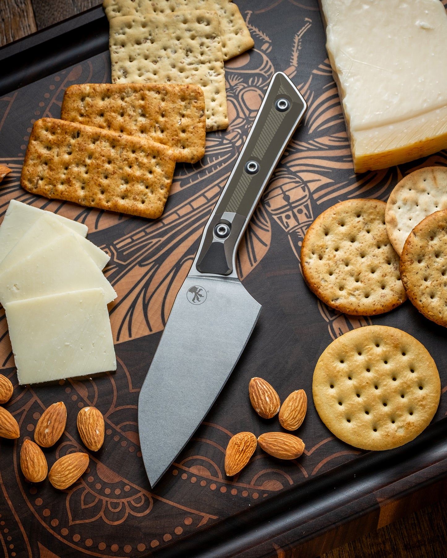 Microtech Culinary Santoku 4.2" Knife with OD green G10 handle, titanium bolsters and stonewashed stainless steel blade on a dragon fly cutting board surrounded by cheeses, crackers and nuts. Premium professional kitchen knife for precision chopping and modern cooking.