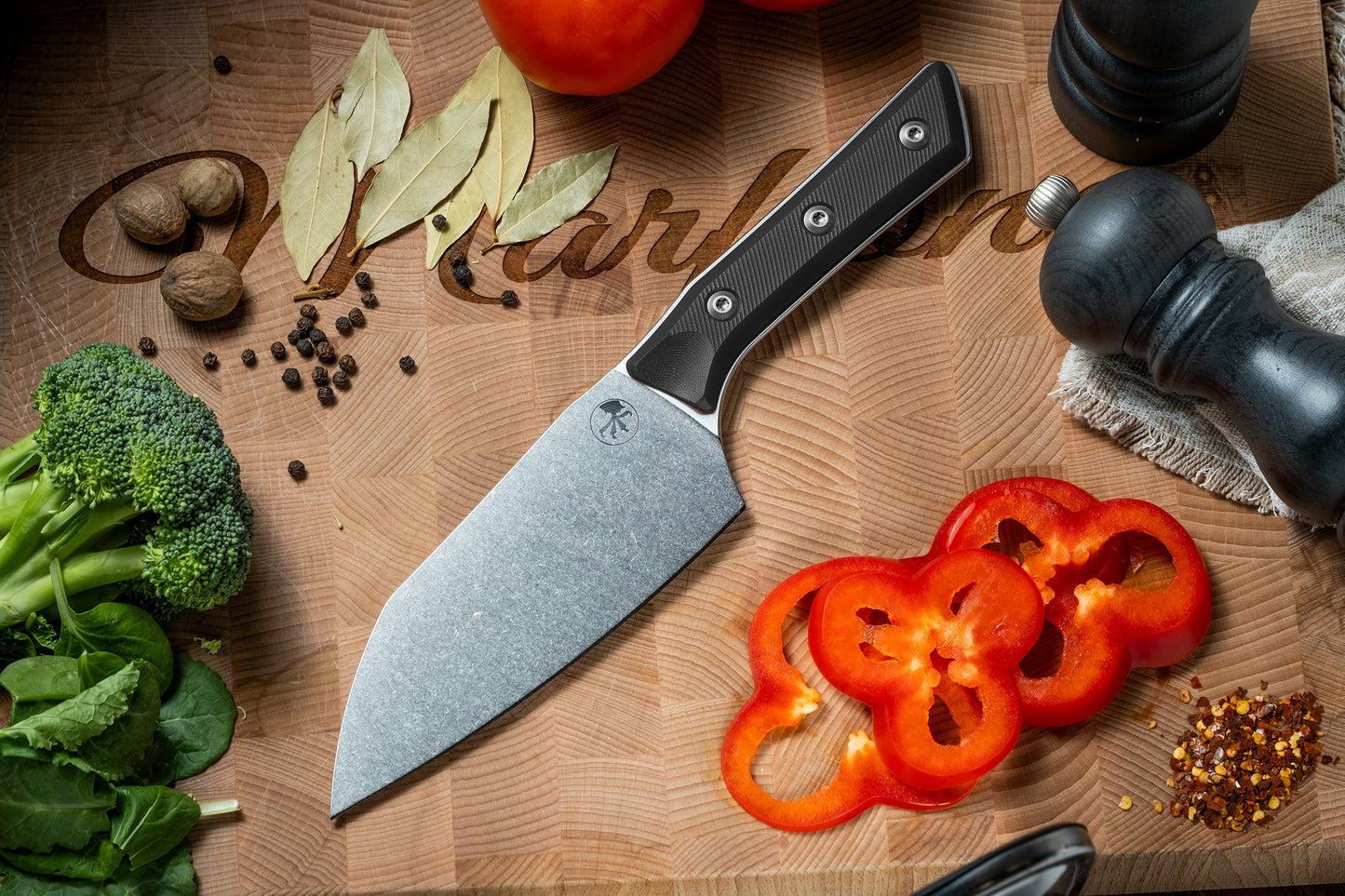 Microtech Culinary Santoku 6" Knife with black G10 handle and stonewashed stainless steel blade on a wooden cutting board surrounded by fresh vegetables and spices. Premium professional kitchen knife for precision chopping and modern cooking.