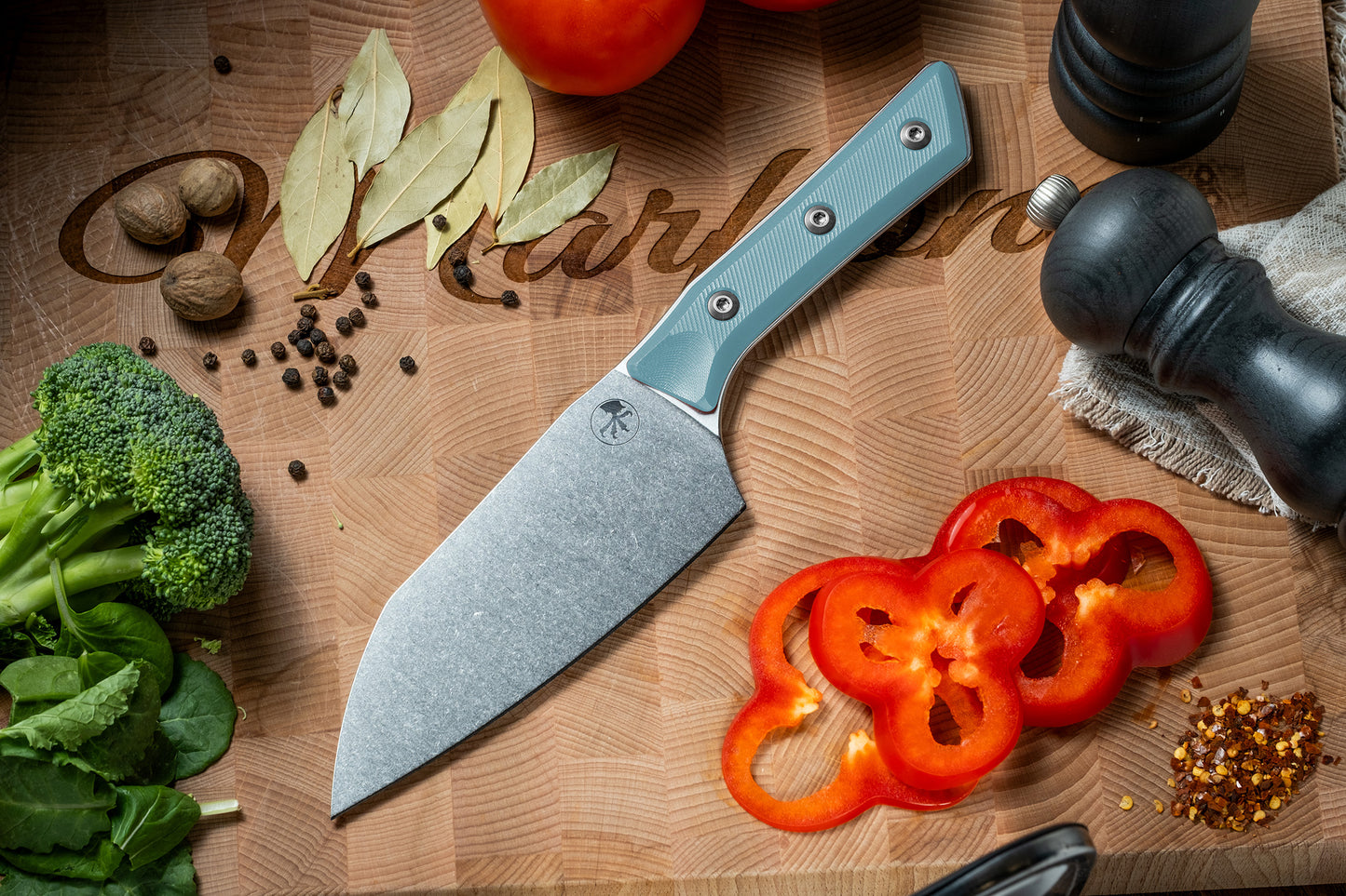 Microtech Culinary Santoku 6" Knife with caribbean blue G10 handle and stonewashed stainless steel blade on a wooden cutting board surrounded by fresh vegetables and spices. Premium professional kitchen knife for precision chopping and modern cooking.