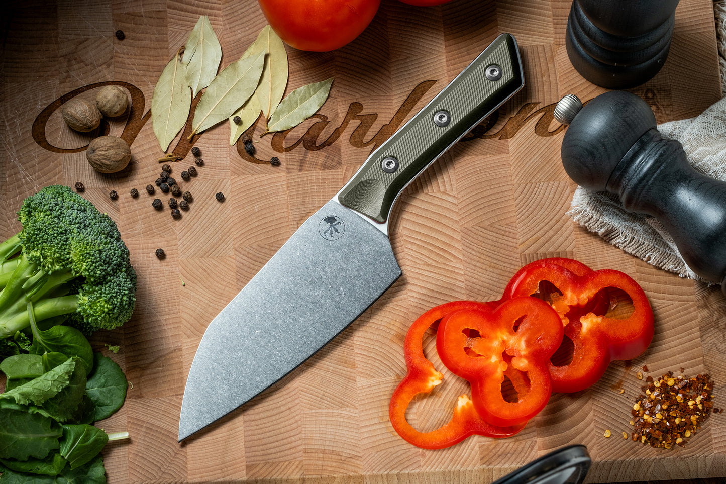 Microtech Culinary Santoku Knife with OD green G10 handle and stonewashed stainless steel blade on a wooden cutting board surrounded by fresh vegetables and spices. Premium professional kitchen knife for precision chopping and modern cooking.