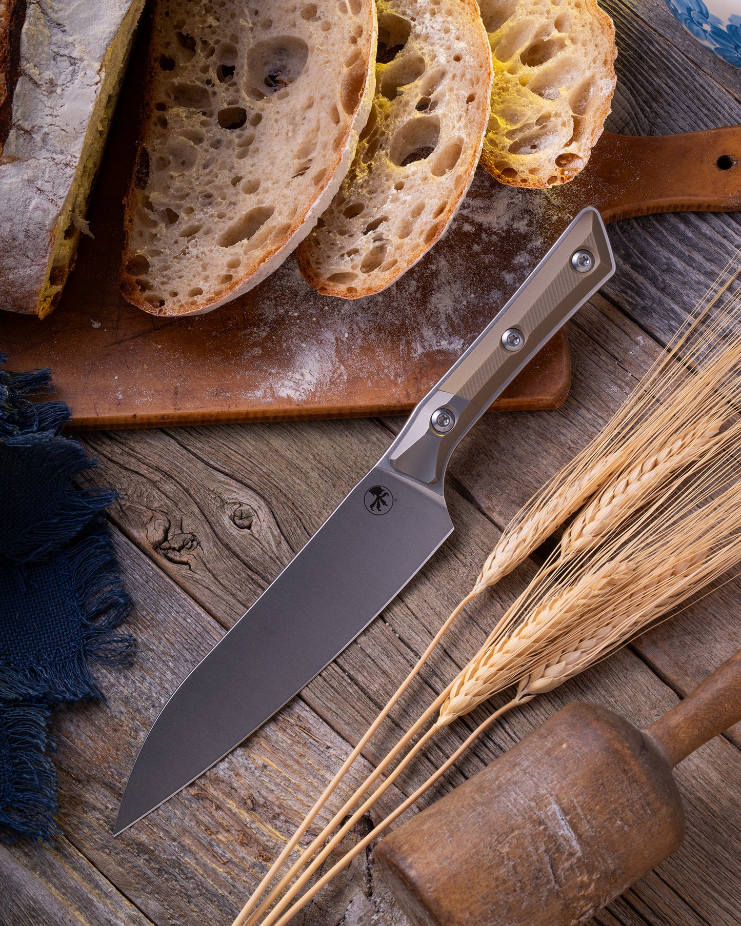 Microtech Culinary Utility Knife with OD green G10 handle, titanium bolsters and stonewashed stainless steel blade on a wooden table surrounded by wheat and freshly cut bread. Premium professional kitchen knife for precision chopping and modern cooking.