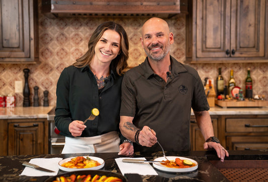 Smiling man and woman in rustic kitchen enjoying plated food, surrounded by vegetables and bottles.