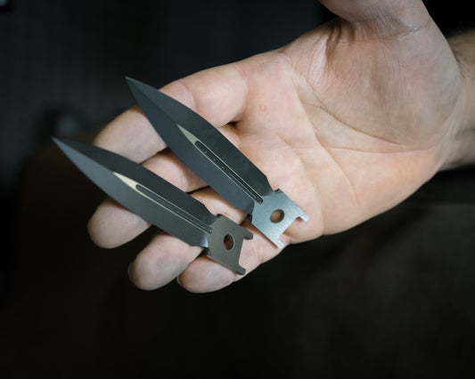 Hand holding two black dagger-shaped steel knife blades with holes near the base