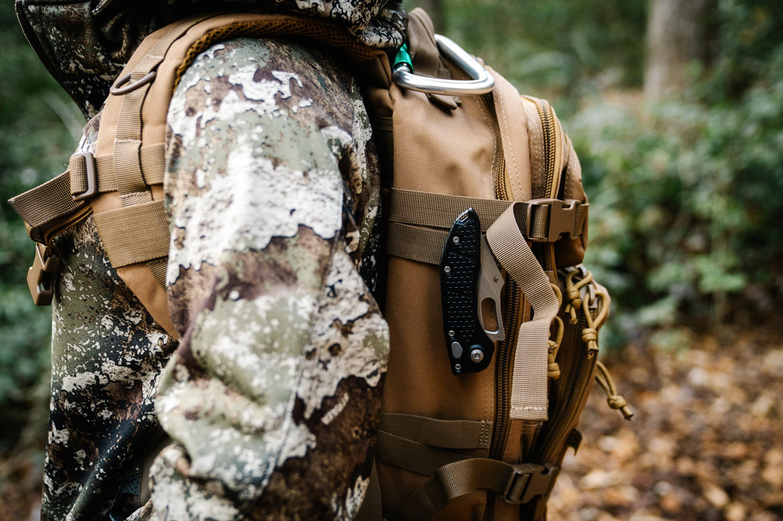 Camouflage jacket and tan tactical backpack with knife in forest camping setting