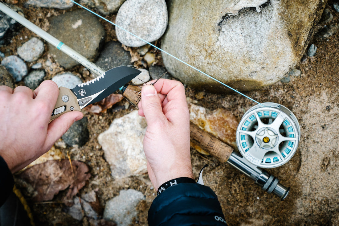 Person preparing fishing line with knife next to fishing reel outdoors on rocks