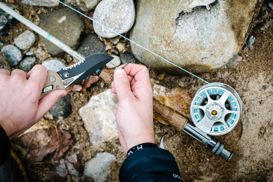 Person preparing fishing line with knife next to fishing reel outdoors on rocks