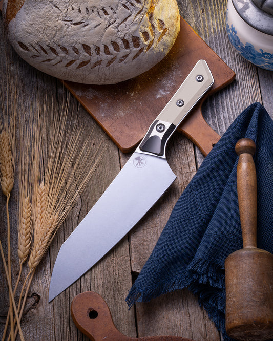 Microtech Culinary Chef Knife featuring an ivory G10 handle and titanium bolster, displayed on a rustic wooden surface beside artisan bread, wheat stalks, and kitchen tools. Premium handcrafted chef knife for professional and home chefs.