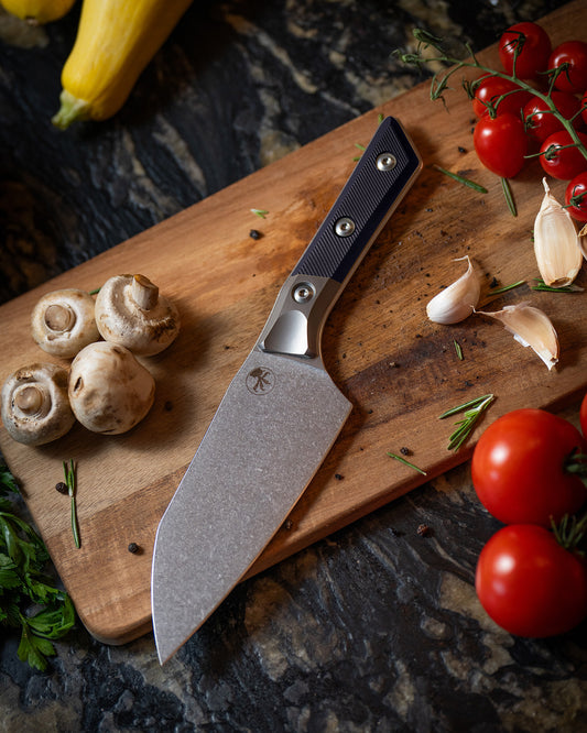 Microtech Culinary Santoku 6" Knife with black G10 handle, titanium bolsters and stonewashed stainless steel blade on a wooden cutting board surrounded by fresh vegetables and spices. Premium professional kitchen knife for precision chopping and modern cooking.