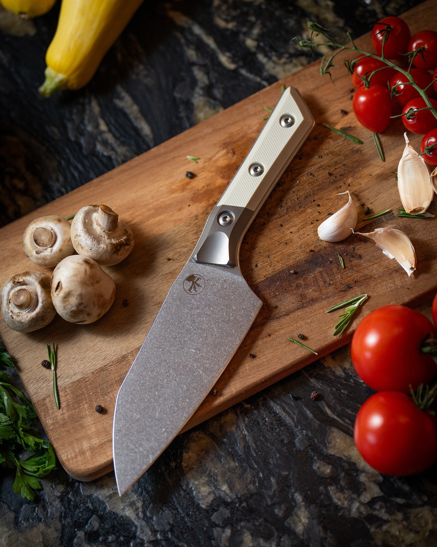 Microtech Culinary Santoku 6" Knife with white G10 handle, titanium bolsters and stonewashed stainless steel blade on a wooden cutting board surrounded by fresh vegetables and spices. Premium professional kitchen knife for precision chopping and modern cooking.