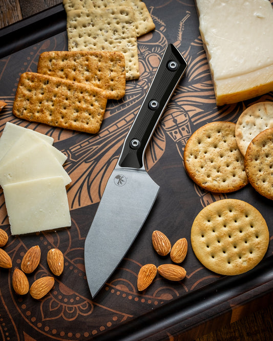 Microtech Culinary Santoku 4.2" Knife with black G10 handle and stonewashed stainless steel blade on a dragon fly cutting board surrounded by cheeses, crackers and nuts. Premium professional kitchen knife for precision chopping and modern cooking.