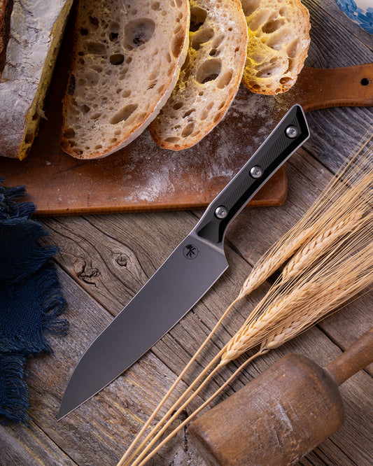 Microtech Culinary Utility Knife with black G10 handle and stonewashed stainless steel blade on a wooden table surrounded by wheat and freshly cut bread. Premium professional kitchen knife for precision chopping and modern cooking.