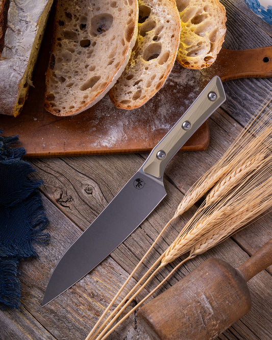 Microtech Culinary Utility Knife with OD green G10 handle and stonewashed stainless steel blade on a wooden table surrounded by wheat and freshly cut bread. Premium professional kitchen knife for precision chopping and modern cooking.