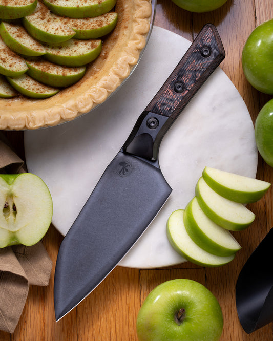 Microtech Culinary Santoku 6" Knife with carbon fiber G10 handle, titanium DLC bolsters and stonewashed stainless steel DLC blade on a wooden cutting board surrounded by fresh apples. Premium professional kitchen knife for precision chopping and modern cooking.