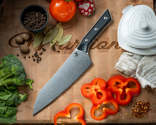 Microtech Culinary Chef Knife with black G10 handle and stonewashed stainless steel blade on a wooden cutting board surrounded by fresh vegetables and spices. Premium professional kitchen knife for precision chopping and modern cooking.