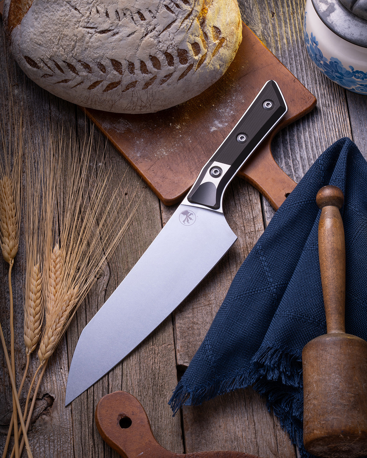 Microtech Culinary Chef Knife featuring a black G10 handle and titanium bolster, displayed on a rustic wooden surface beside artisan bread, wheat stalks, and kitchen tools. Premium handcrafted chef knife for professional and home chefs.