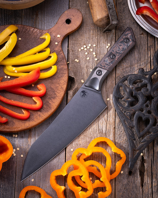Microtech Chef Knife with DLC finish and Livewire Kevlar Carbon Fiber handle featuring Titanium bolster, displayed on rustic cutting board with sliced bell peppers.