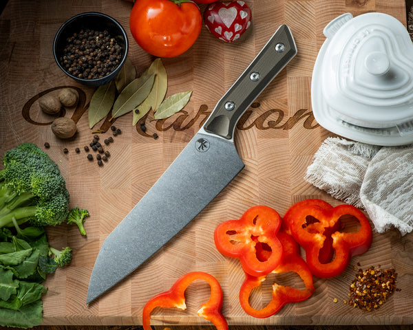 Microtech Culinary Chef Knife with OD Green G10 handle and stonewashed stainless steel blade on a wooden cutting board surrounded by fresh vegetables and spices. Premium professional kitchen knife for precision chopping and modern cooking.