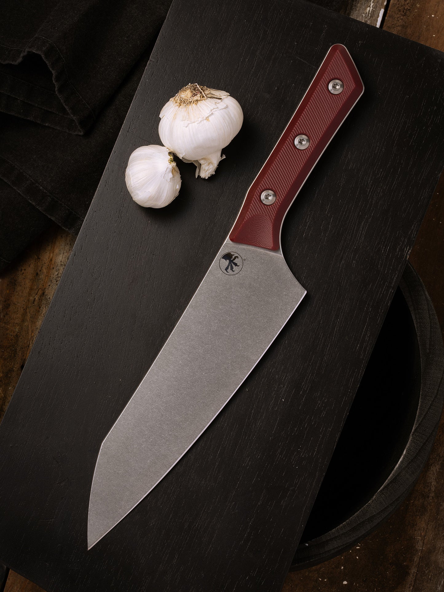 Microtech Knives Culinary Chef Knife in Merlot with a stonewashed stainless steel blade and red textured handle, displayed on a dark wooden cutting board next to fresh garlic bulbs. Premium professional kitchen knife designed for precision cutting and modern culinary performance.