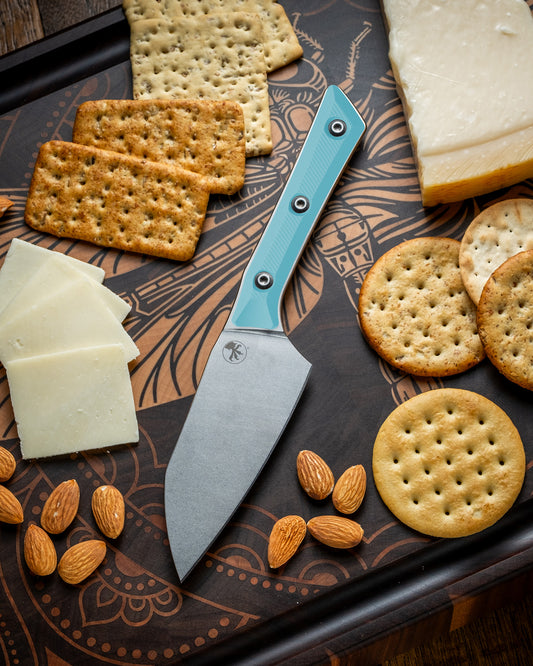 Microtech Culinary Santoku 4.2" Knife with caribbean blue G10 handle and stonewashed stainless steel blade on a dragon fly cutting board surrounded by cheeses, crackers and nuts. Premium professional kitchen knife for precision chopping and modern cooking.