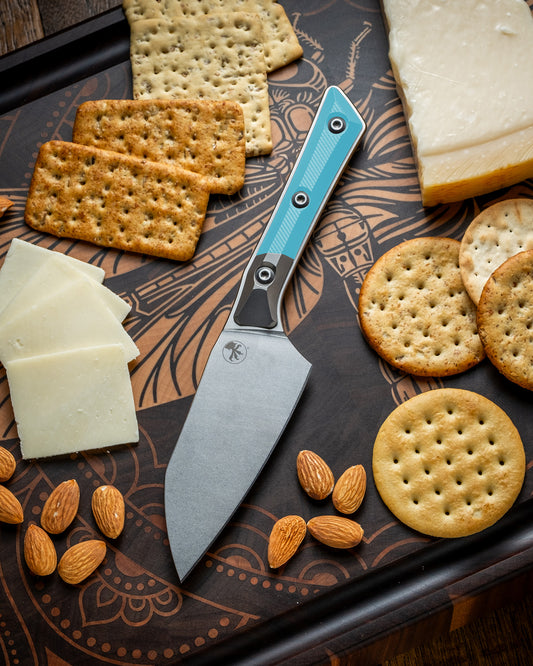Microtech Culinary Santoku 4.2" Knife with caribbean blue G10 handle, titanium bolsters and stonewashed stainless steel blade on a dragon fly cutting board surrounded by cheeses, crackers and nuts. Premium professional kitchen knife for precision chopping and modern cooking.