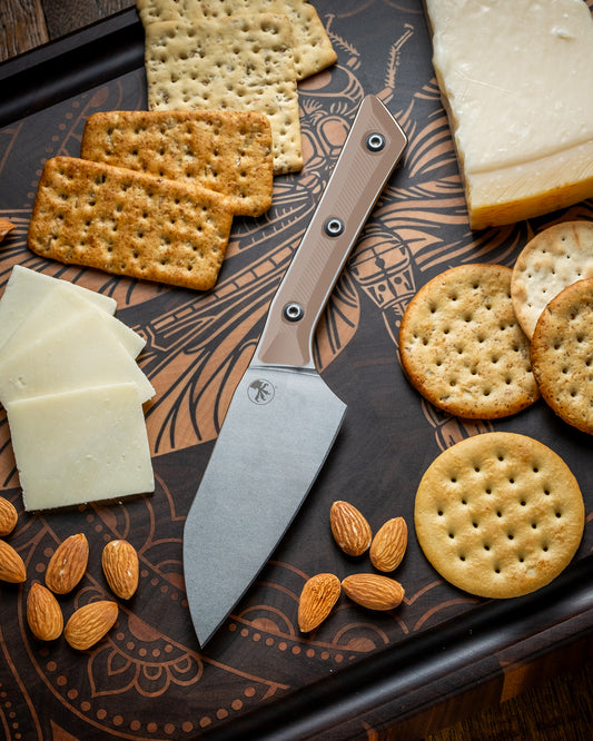 Microtech Culinary Santoku 4.2" Knife with coyote tan G10 handle and stonewashed stainless steel blade on a dragon fly cutting board surrounded by cheeses, crackers and nuts. Premium professional kitchen knife for precision chopping and modern cooking.