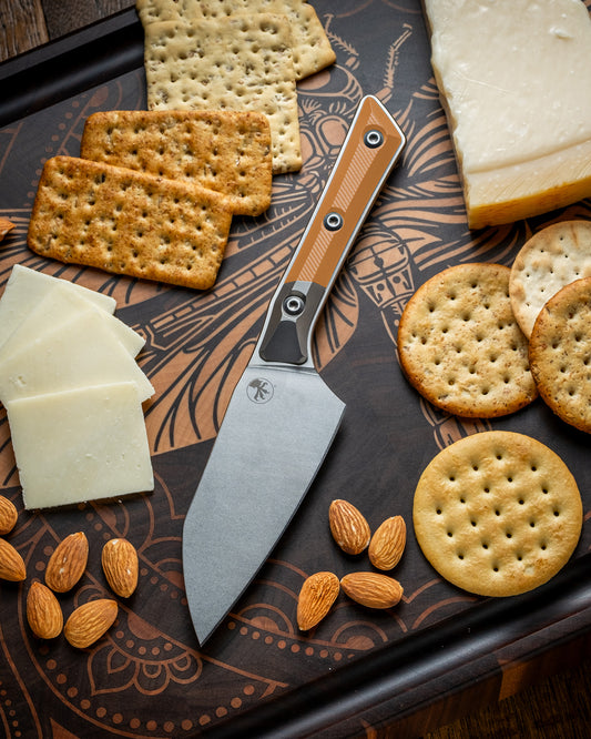 Microtech Culinary Santoku 4.2" Knife with ember G10 handle, titanium bolsters and stonewashed stainless steel blade on a dragon fly cutting board surrounded by cheeses, crackers and nuts. Premium professional kitchen knife for precision chopping and modern cooking.