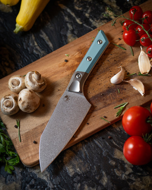 Microtech Culinary Santoku 6" Knife with caribbean blue G10 handle, titanium bolsters and stonewashed stainless steel blade on a wooden cutting board surrounded by fresh vegetables and spices. Premium professional kitchen knife for precision chopping and modern cooking.