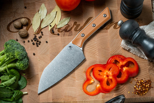 Microtech Culinary Santoku 6" Knife with ember G10 handle and stonewashed stainless steel blade on a wooden cutting board surrounded by fresh vegetables and spices. Premium professional kitchen knife for precision chopping and modern cooking.