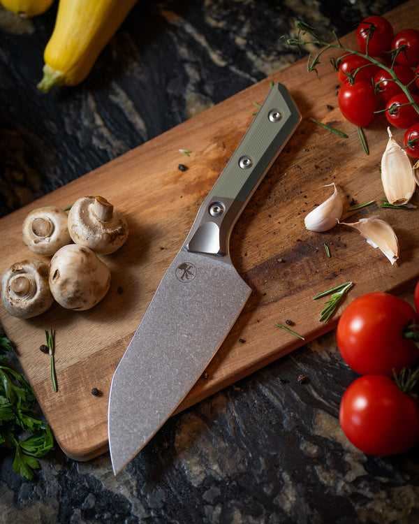 Microtech Culinary Santoku 6" Knife with OD green G10 handle, titanium bolsters and stonewashed stainless steel blade on a wooden cutting board surrounded by fresh vegetables and spices. Premium professional kitchen knife for precision chopping and modern cooking.