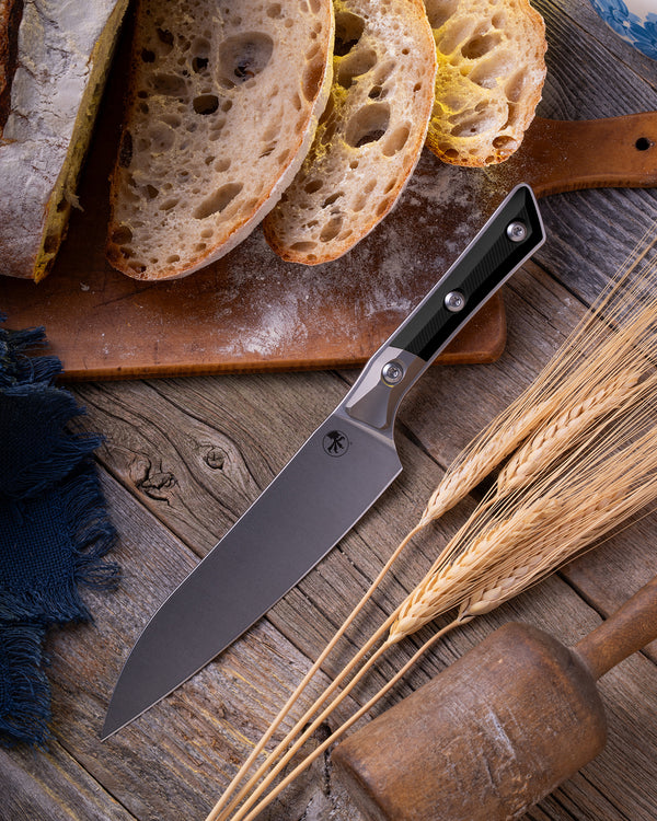 Microtech Culinary Utility Knife with black G10 handle, titanium bolsters and stonewashed stainless steel blade on a wooden table surrounded by wheat and freshly cut bread. Premium professional kitchen knife for precision chopping and modern cooking.