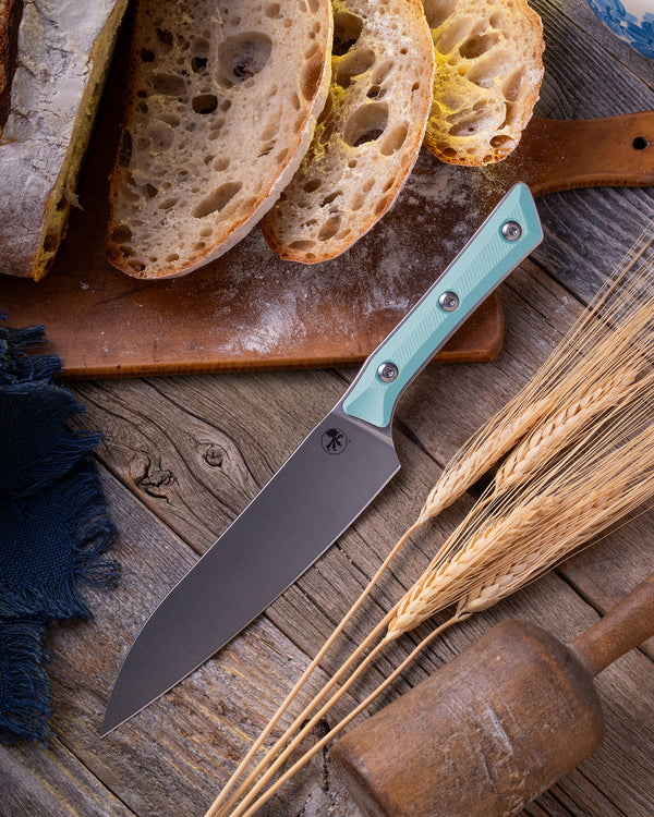 Microtech Culinary Utility Knife with caribbean blue G10 handle and stonewashed stainless steel blade on a wooden table surrounded by wheat and freshly cut bread. Premium professional kitchen knife for precision chopping and modern cooking.