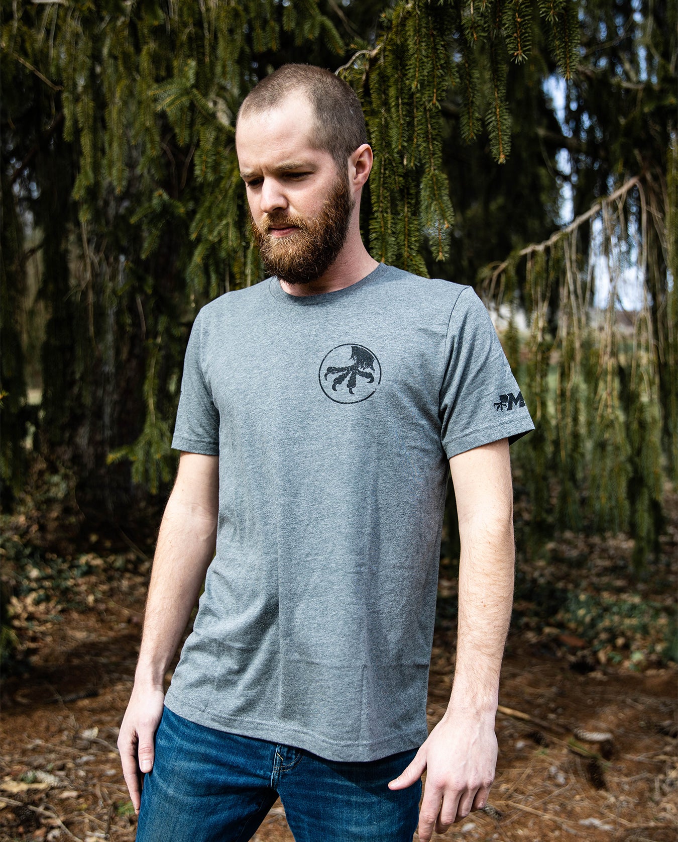 Man wearing gray Bigfoot graphic t-shirt and jeans standing outdoors by pine trees