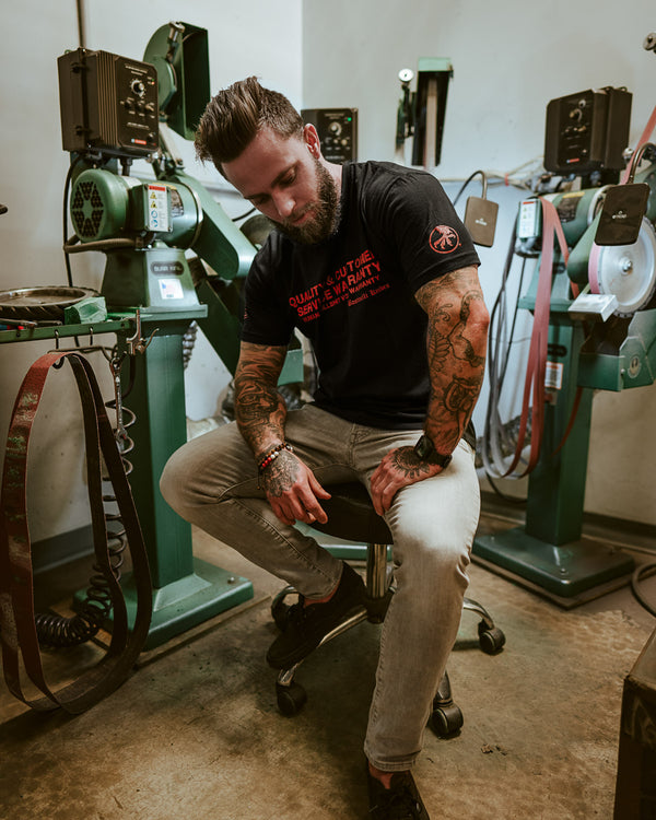 Tattooed man in black graphic t-shirt sitting in industrial workshop with belt sanders